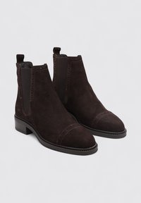 Bottes de cheville en suede marron avec panneaux élastiques sur les côtés, bout arrondi, finitions cousues et petit talon carré. Texture lisse et design classique.