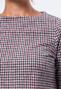 Primer plano de una persona que lleva una camiseta ajustada con un patrón de pata de gallo en burdeos, negro y blanco y un escote redondo.