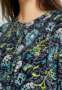 Marinblå blomklänning med ett intrikat mönster i vitt, teal och limegrönt. Slät tyg, knäppdetalj vid halslinningen, avslappnad passform.