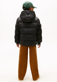 Chaqueta de plumas negra con capucha, con textura acolchada y corte suelto, combinada con pantalones anchos marrones. Una gorra verde completa el conjunto.