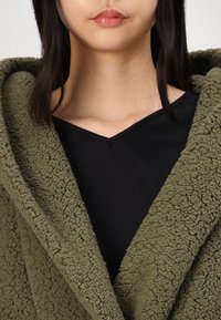 Envuelve de forro polar texturizado en verde oliva, con un cuello chal, superpuesto a una blusa negra de escote en V. La tela parece suave y acogedora, con una textura gruesa y esponjosa.