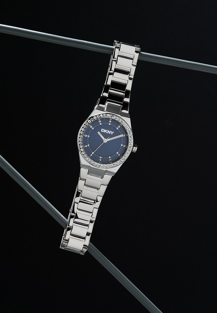 Orologio in metallo argentato con quadrante rotondo blu, lunetta scintillante e design a bracciale. Presenta il logo DKNY nella parte superiore del quadrante.