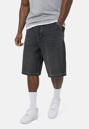 Mężczyzna w czarnych szortach jeansowych, szarej koszulce, białych skarpetkach do kostek i białych trampkach, stojący z jedną ręką w kieszeni.