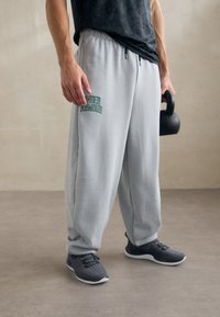 Světle šedé tepláky s elastickým pasem a zeleným logem "Under Armour", doplněné o šedé sportovní boty. Těžká, měkká tkanina.