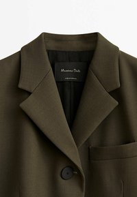 Blazer vert olive foncé cintré avec col cranté, un bouton noir, poche poitrine et étiquette intérieure noire.