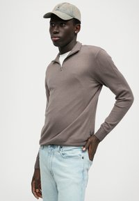 Pruun lukuga kampsun pikkade varrukatega, soonilise äärega, valge särgiga, kerged sinised teksad ja hall müts.