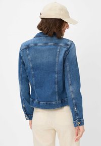 Giacca in denim blu con un design aderente, polsini a bottoni e cuciture sul petto. Presenta una texture leggermente consumata e una silhouette corta.