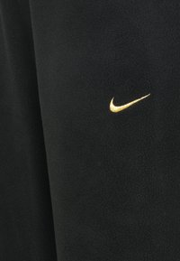 Musta fleece-kangas pehmeällä pinnalla. Edessä on pieni kultainen Nike-logon brodeeraus.