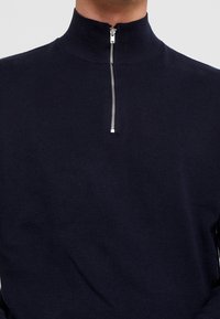 Donkerblauwe ritstrui met hoge kraag, gemaakt van ribgebreid materiaal. Voorzien van een zilveren rits en een aansluitend ontwerp.