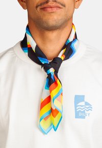 Hombre vestido con una camisa blanca con un logo azul y una bufanda anudada colorida que presenta rayas rojas, amarillas, azules, negras y blancas alrededor de su cuello.