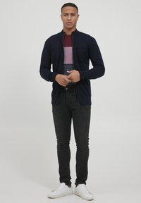 Veste zippée marine avec poches avant, associée à une chemise courte à manches courtes rayée bordeaux, un jean skinny noir et des baskets blanches.