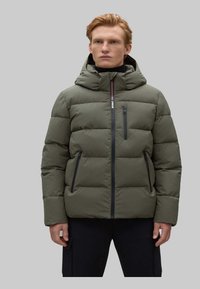 Chaqueta acolchada verde oliva con capucha, con acolchado horizontal, bolsillos con cremallera y cremallera frontal con detalles en contraste.