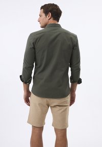 Chemise boutonnée verte foncée avec les manches retroussées, associée à un short beige. Le tissu semble lisse avec une coupe ajustée.
