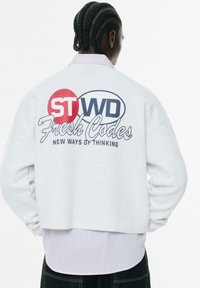 Lys grå cropped sweatshirt med broderet logo i rød og navy, med teksten "Fresh Codes" og "New Ways of Thinking" på ryggen.