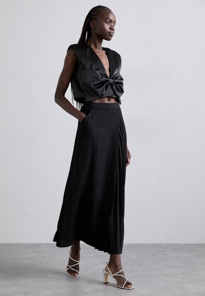 Schwarzes Satin-Crop-Top mit einer geknoteten Vorderseite, kombiniert mit einem langen, plissierten schwarzen Rock. Das Model trägt silberne Riemchenheels.