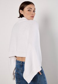 Weißer gestrickter Pullover mit hohem Kragen und Fledermausärmeln, versehen mit gerippten Bündchen und Saumdetails, kombiniert mit hochtaillierten blauen Jeans.