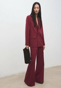 Ensemble constitué d'un blazer sur mesure bordeaux et d'un pantalon évasé, fabriqué en tissu texturé. Le mannequin tient une pochette noire avec un accent doré. Fond simple.