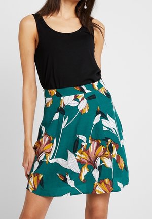 Femme portant un haut noir sans manches et une jupe turquoise avec de grands motifs floraux blancs, jaunes et marron sur un fond uni.