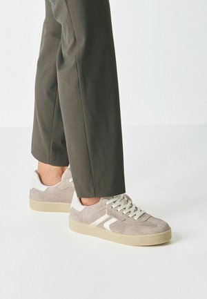 Sneakers in velluto a coste grigio chiaro con dettagli bianchi e lacci, caratterizzate da un motivo strutturato e suola in gomma, abbinate a pantaloni scuri a gamba stretta.