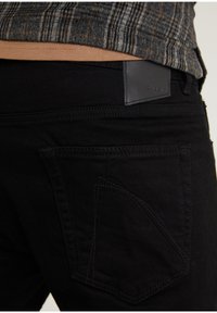Jeans de mezclilla negra con un tejido texturizado, que presentan un diseño de bolsillo distintivo y una etiqueta de cuero negro mate en la cintura.