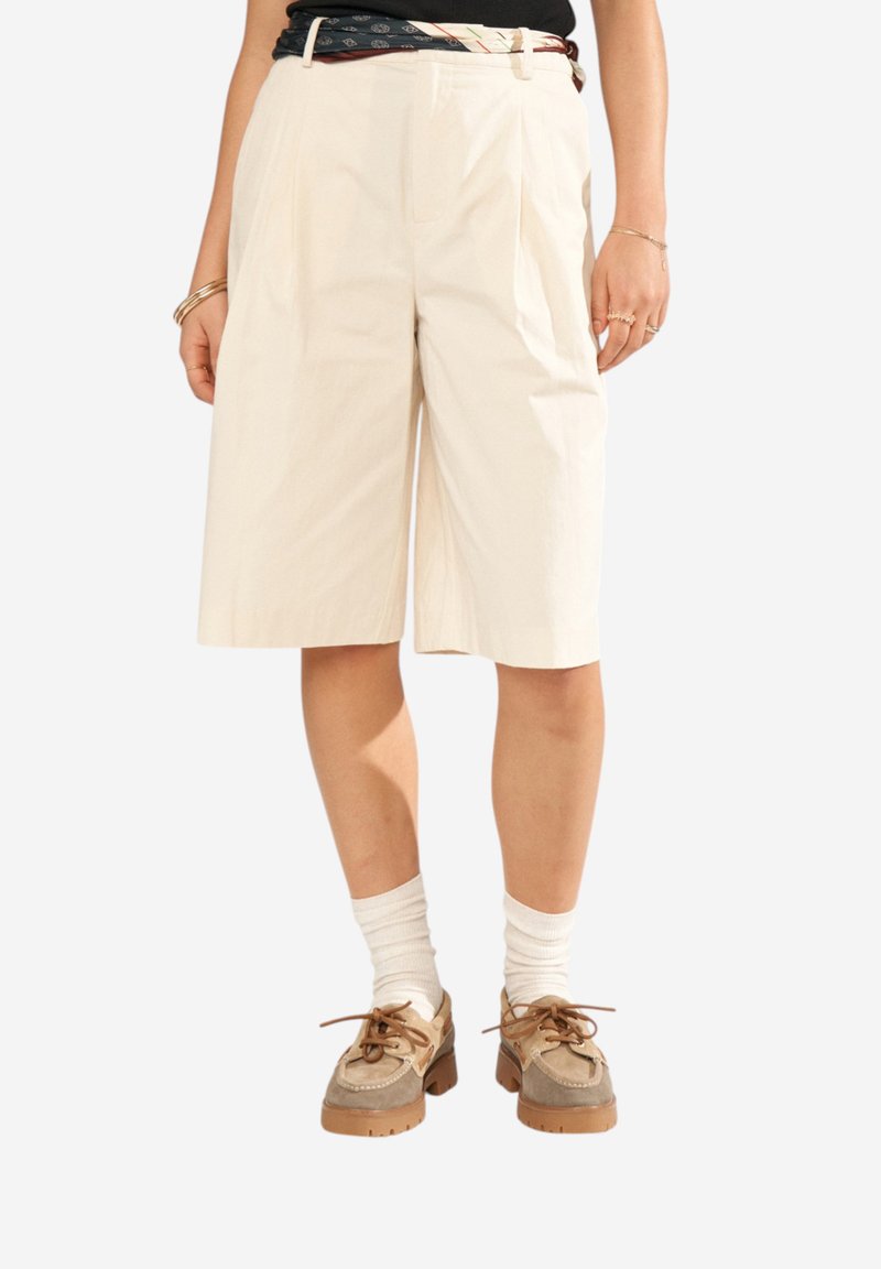 Personne portant un short beige au genou, des chaussettes blanches, des chaussures marron à lacets, un haut noir et une ceinture à motifs, debout les mains détendues.