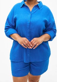 Camicia blu a maniche lunghe con texture e maniche arrotolate abbinata a pantaloni corti blu coordinati. Entrambi hanno una vestibilità ampia e un sottile motivo tessuto.