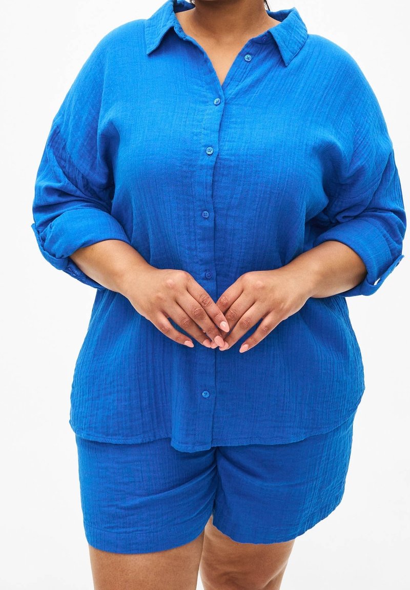 Camicia blu a maniche lunghe con texture e maniche arrotolate abbinata a pantaloni corti blu coordinati. Entrambi hanno una vestibilità ampia e un sottile motivo tessuto.