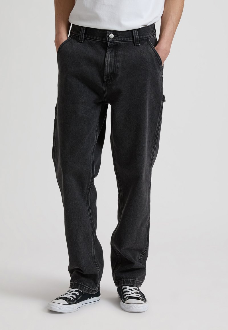 Άτομο που φορά χαλαρά μαύρα τζιν και μαύρα χαμηλά αθλητικά παπούτσια με λευκά κορδόνια, στέκεται με τα χέρια στις τσέπες σε απλό φόντο.