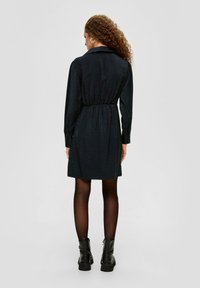 Schwarzes Hemdkleid mit langen Ärmeln, elastischem Bund und Kragen. Getragen mit schwarzen Ankle Boots und durchsichtigen schwarzen Strumpfhosen. Glattes, strukturiertes Material.