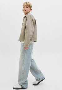 Veste en denim beige clair avec deux poches poitrine, portée sur une chemise blanche ; associée à un jean bleu clair ample et des baskets blanches.