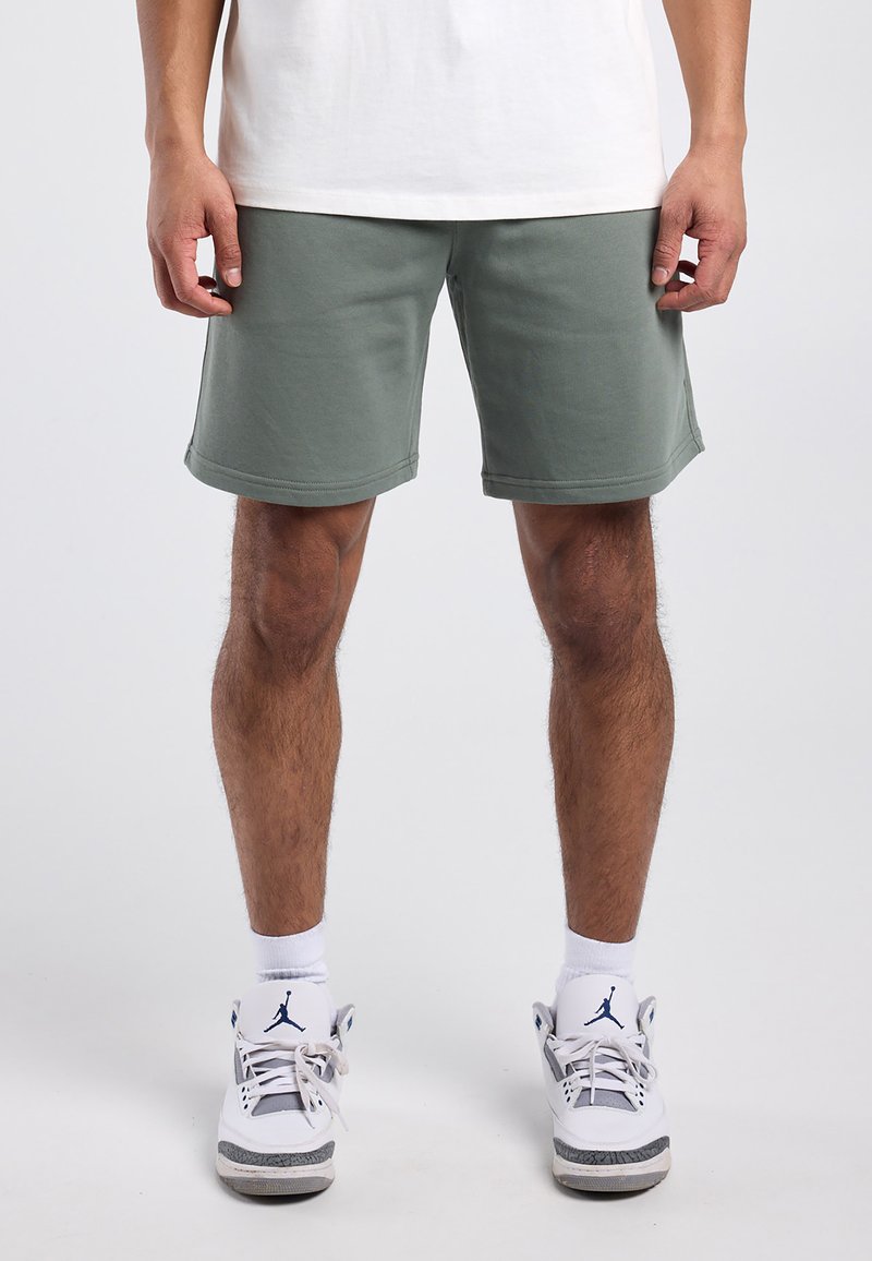 Mann trägt graue kniehohe Shorts, weiße Sportsocken und weiße Turnschuhe mit blauem Logo, steht vor einem schlichten hellen Hintergrund.