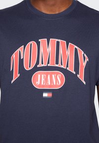 T-shirt de algodão azul-marinho com texto ousado em vermelho e branco "TOMMY JEANS", com um pequeno logótipo em vermelho, branco e azul abaixo do texto.