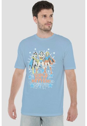 Mann trägt hellblaues T-Shirt mit Comic-Stil weiblichen Superheldinnen und Text "Girls Do It Better" vorne, stehend vor einfarbigem Hintergrund.