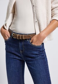 Cinturón de ante marrón con herrajes plateados, usado sobre unos pantalones de mezclilla azul oscuro de textura suave, combinado con una blusa crema debajo de una chaqueta esponjosa crema.