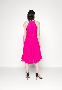 Abito halter fucsia con gonna plissettata, vita arricciata e dettaglio a chiave sulla schiena. Indossato con sandali neri con tacco alto.