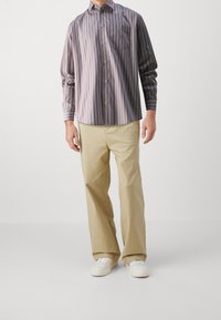 Camicia a righe con maniche lunghe in tonalità scure e chiare, abbinata a pantaloni beige e sneakers bianche. Presenta una tasca sul petto.