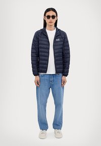 Doudoune marine avec des coutures horizontales, capuche et logo EA7. Portée par-dessus un t-shirt blanc, associée à un jean bleu clair et des chaussures blanches.
