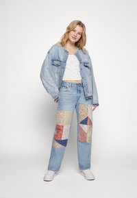 Lichtblauwe spijkerjack gecombineerd met een wit knopen-crop top en blauwe jeans met kleurrijke patchworkpanelen in verschillende patronen.