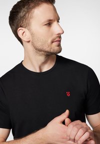 Czarna koszulka z krótkim rękawem wykonana z gładkiego materiału, z małym czerwonym logo na lewej piersi. Prosty okrągły dekolt bez zdobień.