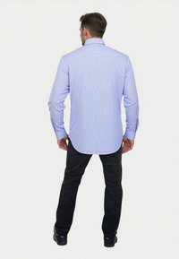 Uomo di spalle che indossa una camicia a righe blu chiaro, pantaloni neri e scarpe scure su uno sfondo chiaro e semplice.