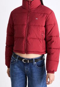 Przycięta czerwona kurtka puchowa z zamkiem i małym logo, noszona z niebieskimi dżinsami i czarnym paskiem, odsłaniająca brzuch i ręce w luźnej pozycji przy bokach.