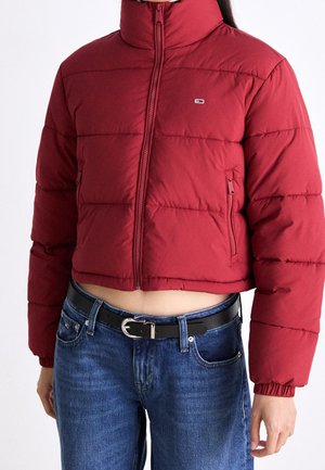 Afskåret rød pufferjakke med lynlås og lille logo, båret med blå jeans og sort bælte, som viser maven og hænderne afslappet ved siderne.