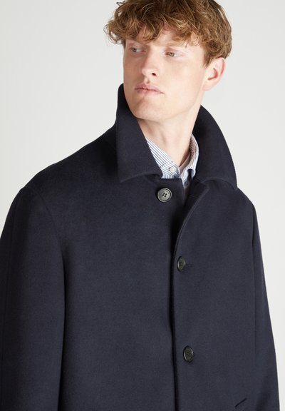 Uomo con capelli ricci che indossa un cappotto di lana blu scuro con colletto alzato e una camicia con strisce sotto.