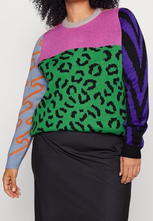 Frau trägt einen mehrfarbigen Pullover mit Tiermuster und abstrakten Designs, kombiniert mit einem schwarzen Rock, gezeigt vom Hals bis zu den Hüften.