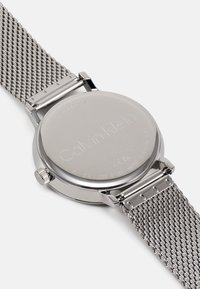 Montre en acier inoxydable avec un bracelet en maille, boîtier rond, finition métallique et fond gravé avec le logo de la marque et des détails sur la résistance à l'eau.