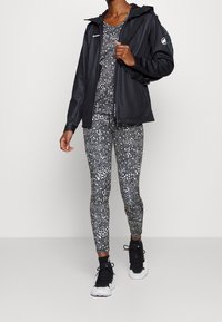 Giacca nera impermeabile, leggings a fantasia con macchie bianche astratte, sneakers nere e uno zaino nero. Design semplice e tessuto leggero.