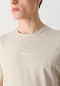 Κοντινό πλάνο ενός ατόμου που φοράει μπεζ κοντομάνικη μπλούζα με την επιγραφή "BOSS" σε λευκό χρώμα στο στήθος.