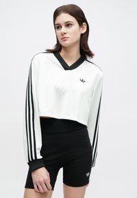 Moteris vilkinti balto ilgo kuriečio medvilninį marškinėlį su juodais dryžiais ir juodus aukšto juosmens šortus, abiejose matosi Adidas logotipai.