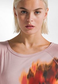 Helles rosa Shirt in lässiger Passform mit weitem Ausschnitt, das auf der Vorderseite einen bunten abstrakten Druck in Orange- und Rottönen zeigt. Weicher Stoff.