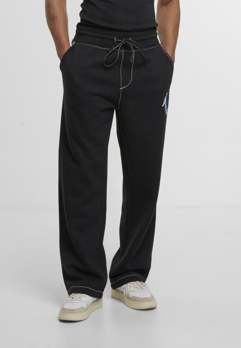 Pantalones de chándal negros con cintura ajustable, costuras blancas en contraste, bolsillos laterales y un detalle gráfico azul en la parte inferior de la pierna izquierda.